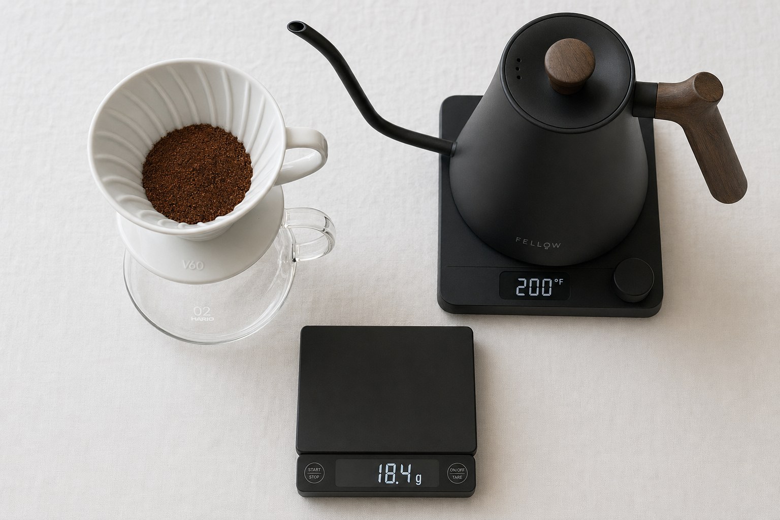Hario V60 ceramic dripper on a glass server next to a Fellow Stagg EKG gooseneck kettle, with a digital scale showing 18.4 g coffee